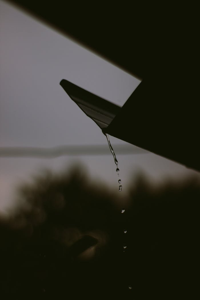 Silhouetted raindrops cascading from a roof edge, set against a moody sky.